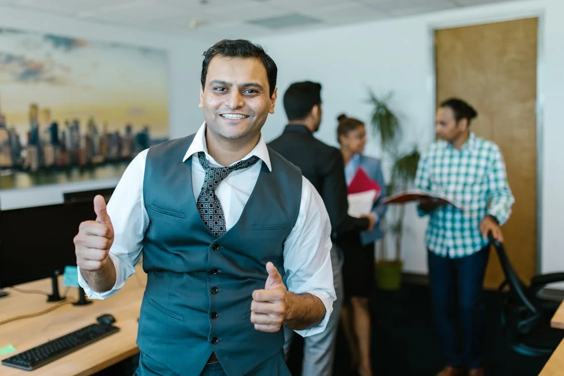 Man standing in an office giving a happy thumbs-up for group health insurance, with employees chatting behind him.
